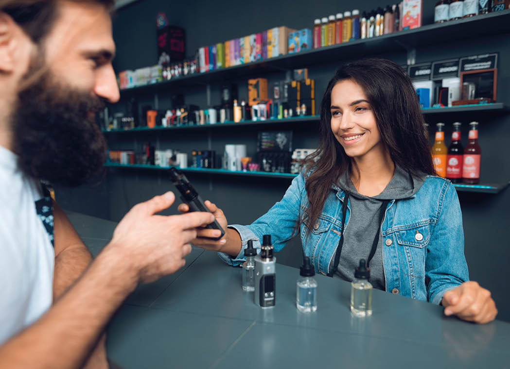 Girl seller shows the choice of electronic cigarettes in vapeshop. Nearby is a buyer - a man with a beard. The store has a large assortment of electronic cigarettes.