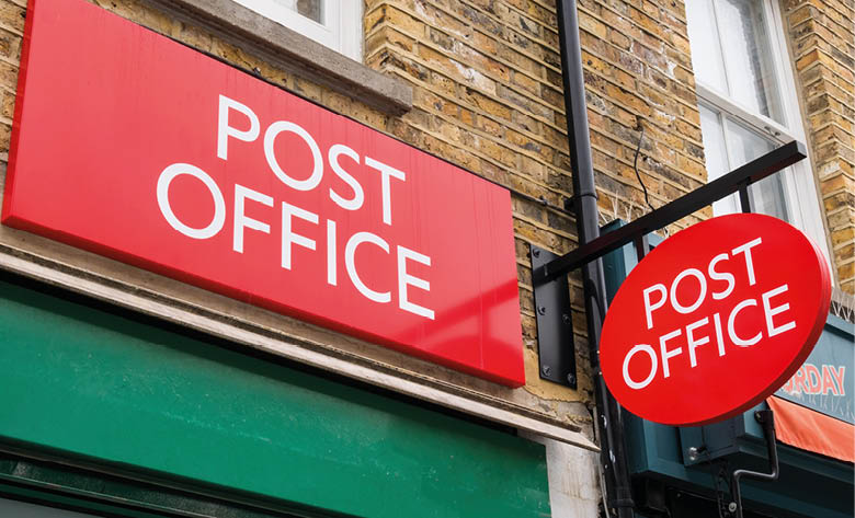 A brick building features two red signs with white text reading “POST OFFICE" near two windows. Description generated by AI