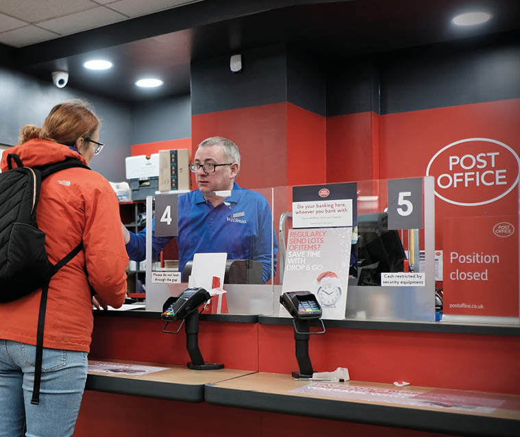 Two adults stand at a post office counter speaking with a clerk behind a protective screen, with one adult wearing a red jacket and backpack. Description generated by AI