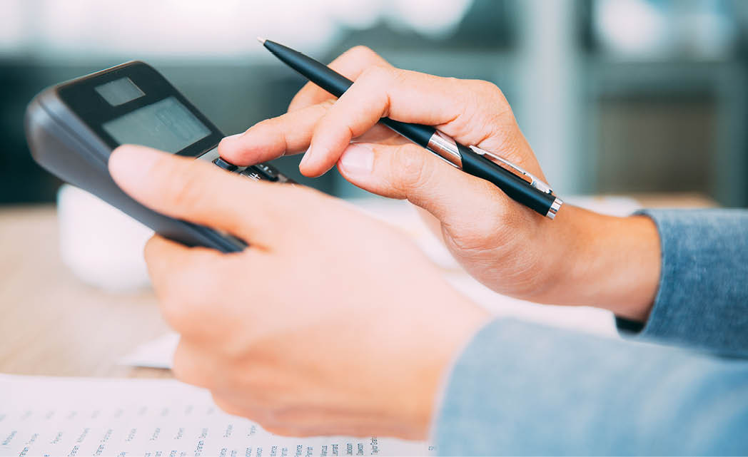 A person wearing a blue long-sleeve shirt is using a calculator with one hand while holding a pen in the other hand over a table with papers. Description generated by AI