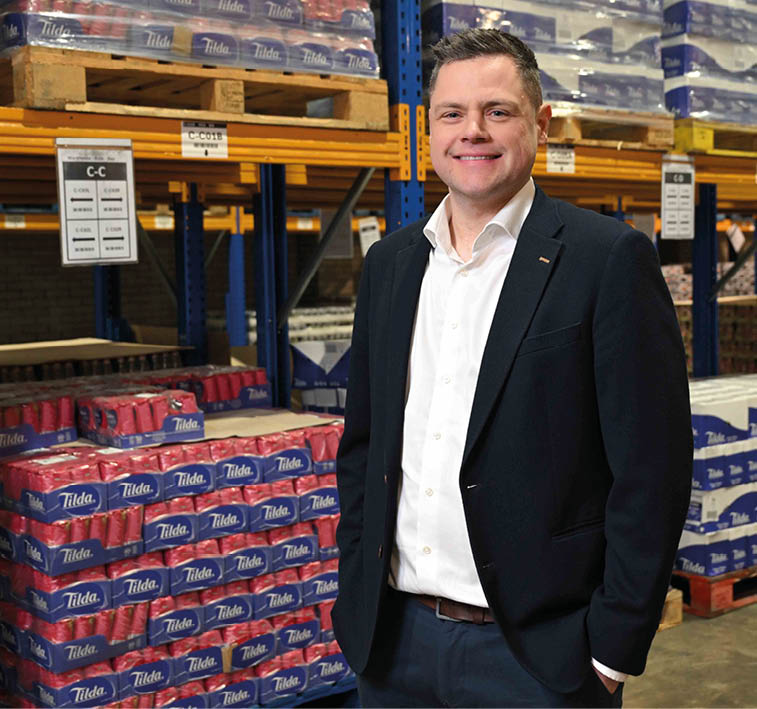 A person wearing a dark suit and white shirt stands in a warehouse aisle with pallets of stacked packaged goods in the background. Description generated by AI