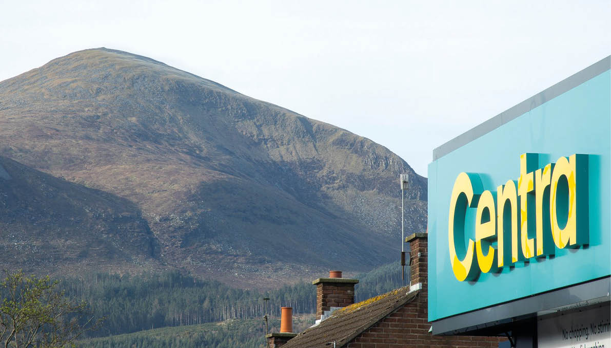 A large mountain rises behind a row of rooftops with chimneys and a partial view of a blue and yellow Centra store sign. Description generated by AI
