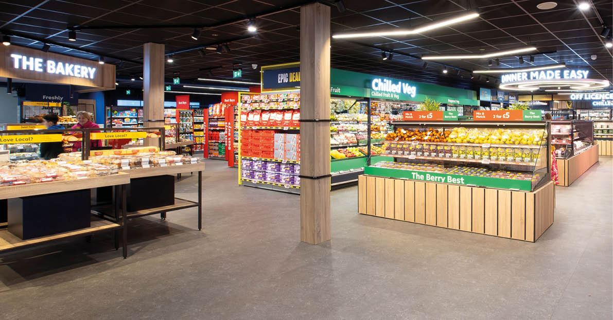 A brightly lit supermarket aisle displays various baked goods, snacks, and fresh produce with clear signage above each section. Description generated by AI