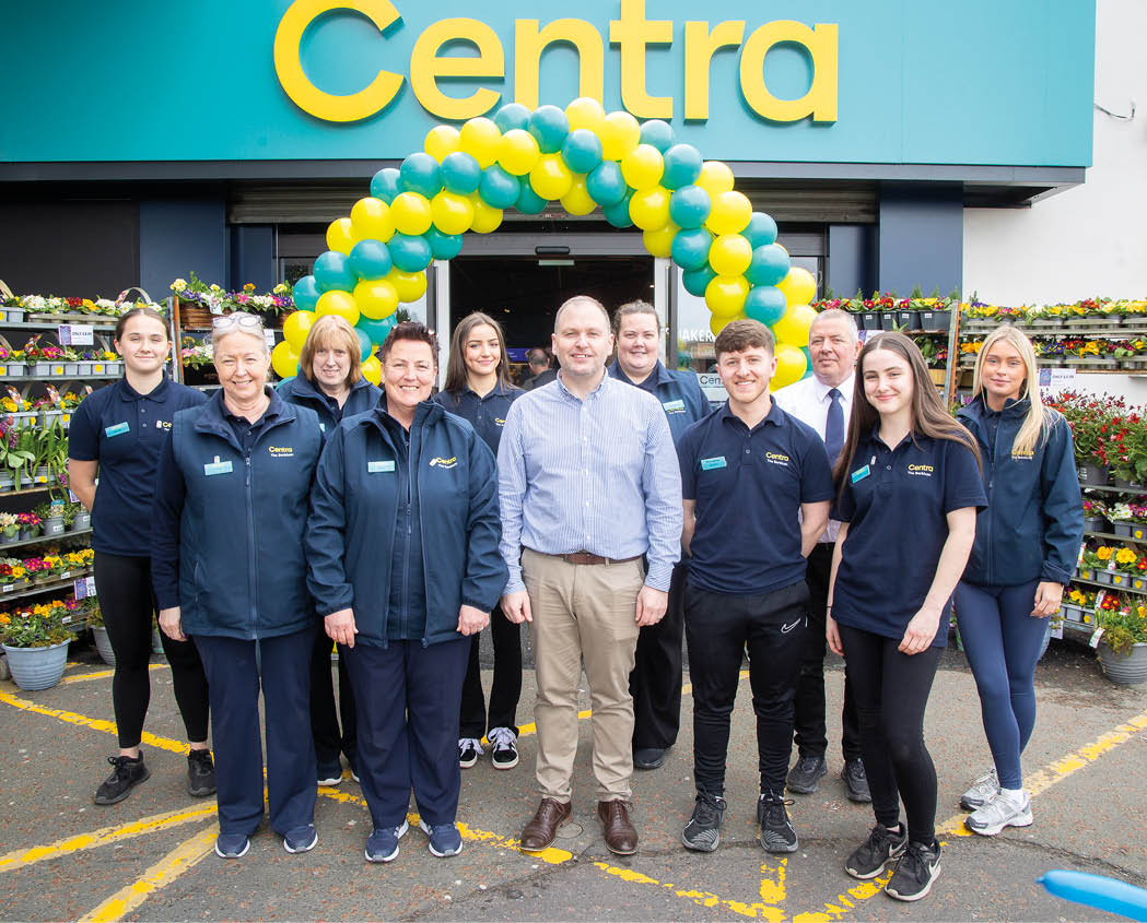 A group of people wearing casual and uniform clothing stand smiling in front of a store entrance decorated with a yellow and green balloon arch and surrounded by racks of flowers. Description generated by AI