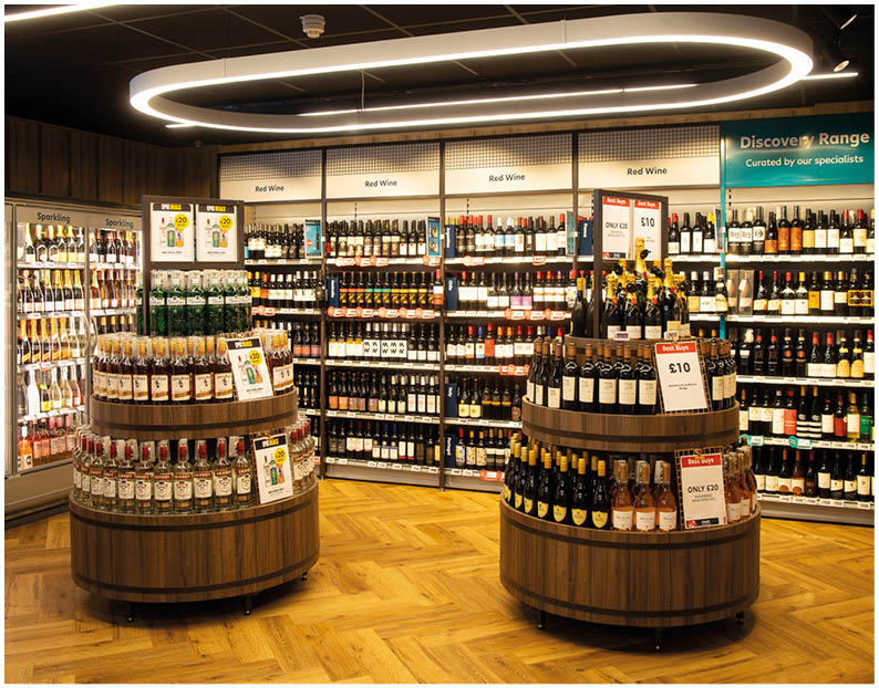 A well-lit wine store interior with wooden floors, circular display tables filled with wine bottles, and shelves stocked with various wines along the walls. Description generated by AI