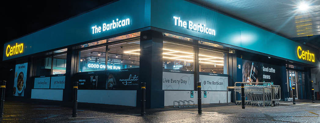 A brightly lit convenience store named Centra and The Barbican is shown at night with wet pavement reflecting the lights. Description generated by AI