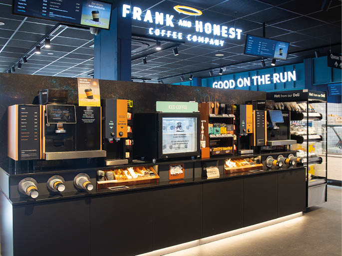 A modern self-service coffee station with multiple coffee machines, cup dispensers, and a display screen in a well-lit convenience store setting. Description generated by AI