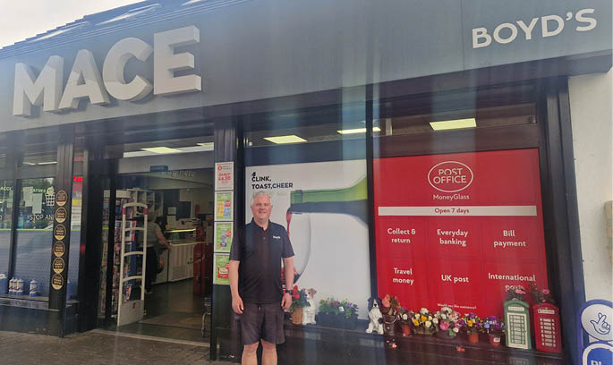 A person wearing a dark shirt and shorts stands in front of a MACE and Boyd's store with a red post office sign and various items displayed in the window. Description generated by AI