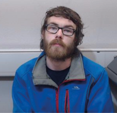A person with glasses and a beard is sitting indoors wearing a blue jacket with red zippers. Description generated by AI