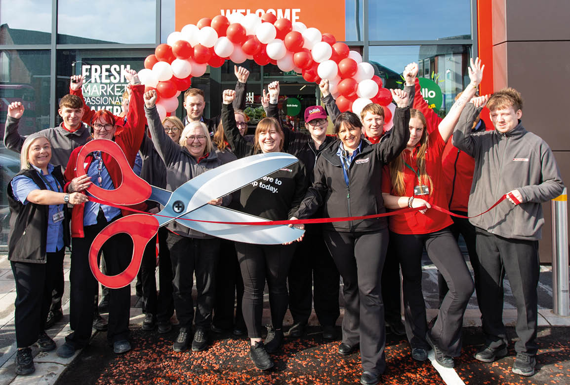 A group of people standing outside a store under a red and white balloon arch are celebrating a ribbon-cutting ceremony with a large pair of scissors. Description generated by AI
