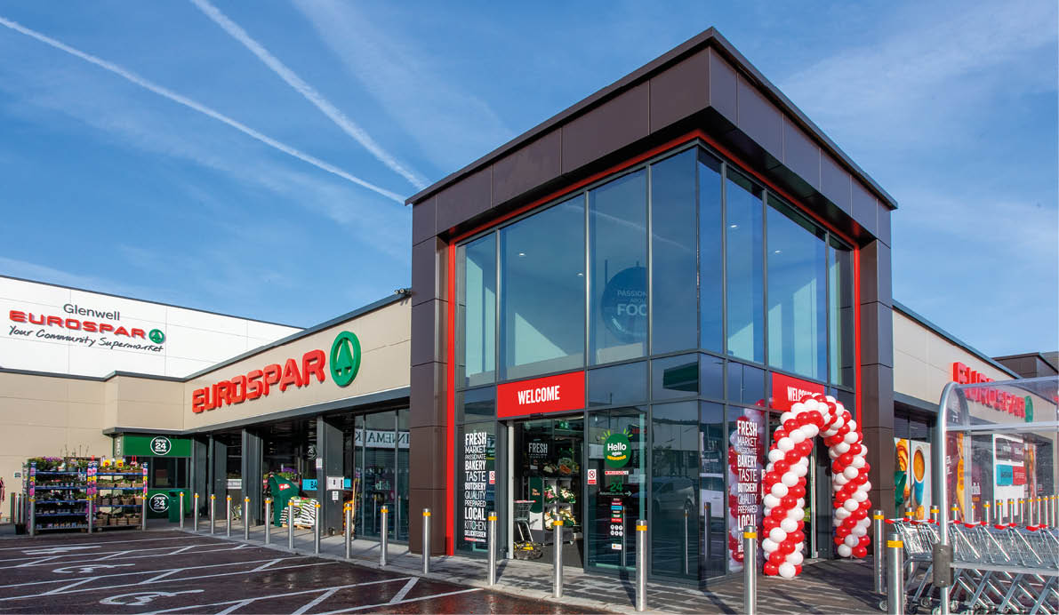 A modern Eurospar supermarket entrance decorated with a red and white balloon arch and a wet parking lot with marked spaces including a handicap spot. Description generated by AI