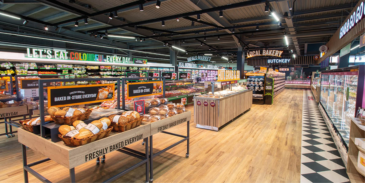 A brightly lit supermarket aisle features wooden displays filled with freshly baked bread and pastries, with signs promoting fresh and colorful food. Description generated by AI