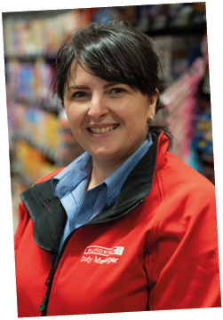 A person with short dark hair wearing a red jacket labeled Duty Manager and a blue collared shirt is smiling in a store aisle. Description generated by AI