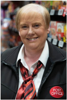 A person with short light hair is smiling and wearing a black jacket with a red and black striped scarf and a white collared shirt, standing in a store aisle. Description generated by AI