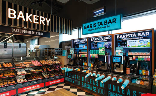 A brightly lit bakery and barista station features shelves filled with assorted pastries and three coffee machines under illuminated signs reading “BAKERY" and "BARISTA BAR." Description generated by AI