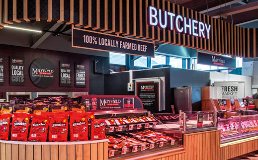 A brightly lit butcher shop display features wooden counters with packaged locally farmed beef products and a neon sign reading “BUTCHERY" above. Description generated by AI