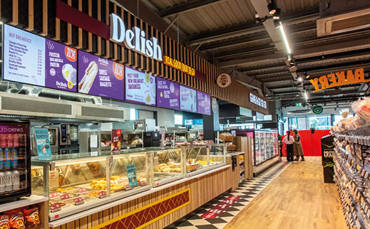 A brightly lit deli counter inside a store displays various baked goods and snacks with digital menu boards above and people standing near the entrance in the background. Description generated by AI