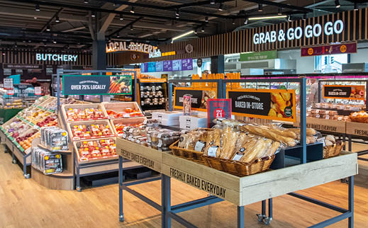 A brightly lit grocery store interior with wooden floors features various food displays including fresh produce, baked goods, and packaged items, with several people shopping in the background. Description generated by AI