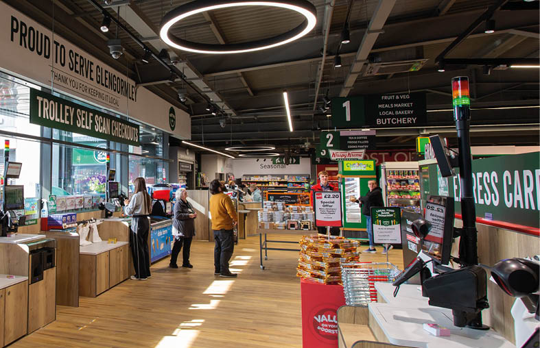 A modern grocery store interior with wooden floors, self-checkout stations, and several people shopping near the produce and bakery sections. Description generated by AI