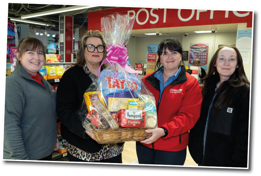 Four adults standing indoors in front of a post office counter holding a gift basket wrapped in clear plastic with a pink bow. Description generated by AI