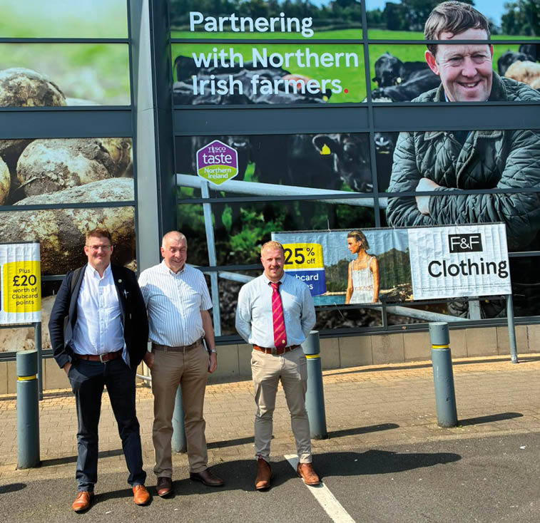 Three adults stand outside a building with large windows displaying advertisements for partnering with Northern Irish farmers, including images of potatoes, cows, and clothing promotions. Description generated by AI