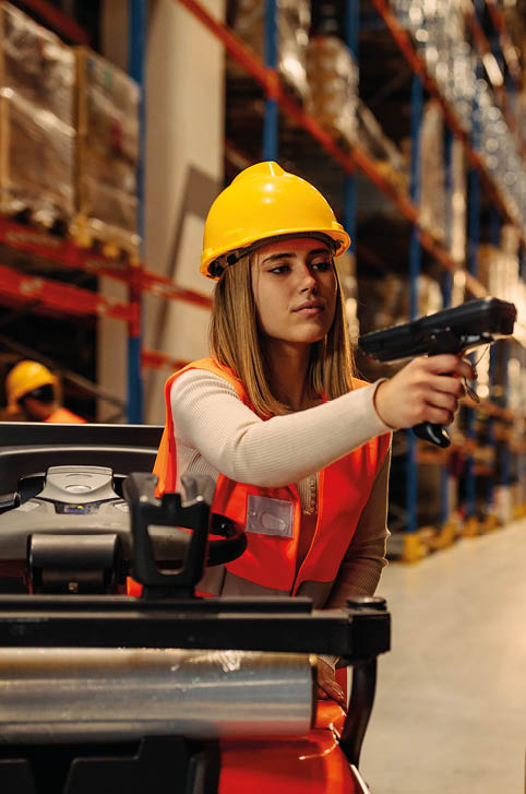 Female warehouse worker with safety helmet is working with bar code scanner while sitting on forklift in warehouse