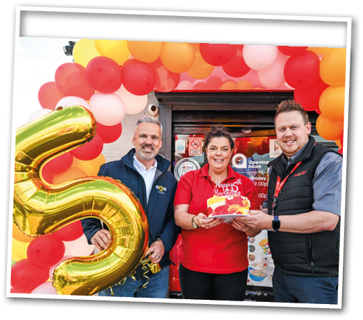 Three adults stand smiling under a colorful balloon arch while holding a birthday cake and a large gold balloon shaped like the number five. Description generated by AI