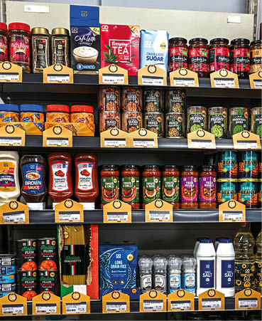 A store shelf stocked with various condiments, sauces, spices, and beverages arranged in rows with clear labels and price tags. Description generated by AI