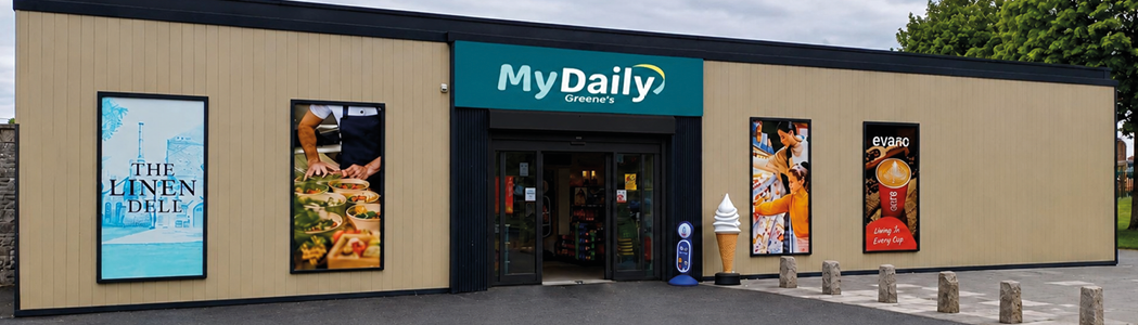 A modern convenience store named MyDaily with promotional posters for food and beverages displayed on the exterior walls and a large paved area in front. Description generated by AI