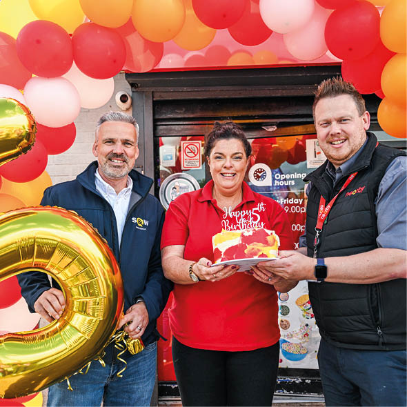 Three adults stand smiling under a colorful balloon arch while holding a birthday cake and a large gold balloon shaped like the number five. Description generated by AI