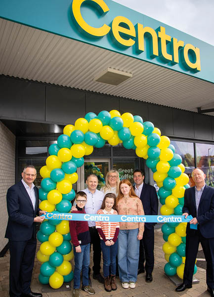 A group of people stands smiling under a yellow and green balloon arch holding a blue ribbon with the word Centra in front of a Centra store during a ribbon-cutting event. Description generated by AI
