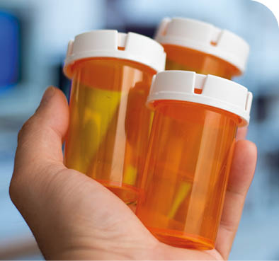 A hand is holding three orange prescription pill bottles with white caps against a blurred background. Description generated by AI
