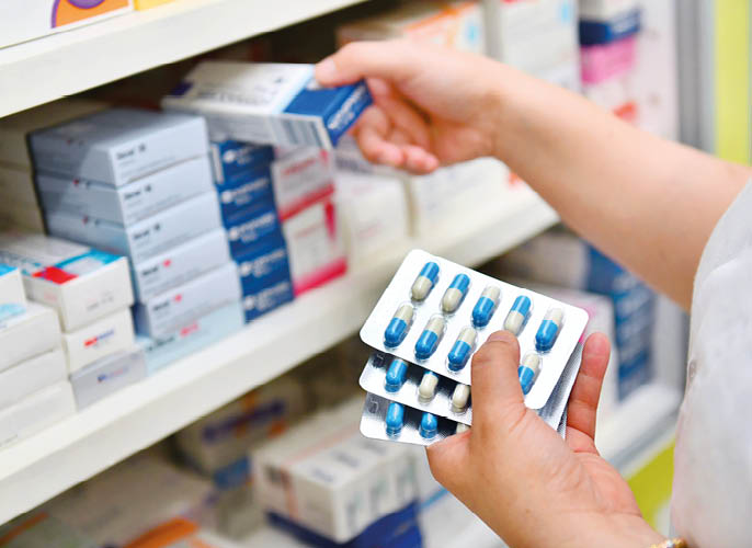 A person is holding blister packs of blue and white capsules while selecting a medication box from a shelf filled with various pharmaceutical products. Description generated by AI
