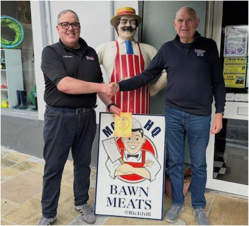 Two adults wearing casual clothing are shaking hands and standing on either side of a butcher shop sign with a cartoon butcher figure and a life-sized statue of a butcher in an apron and hat behind them. Description generated by AI