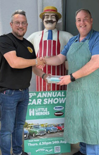 Two adults smiling and shaking hands in front of a colorful statue of a person wearing a hat and apron, with a sign advertising an annual car show visible behind them. Description generated by AI