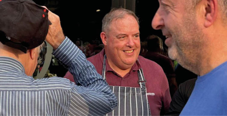 Three adults are engaged in conversation outside, with one wearing a striped apron and smiling. Description generated by AI