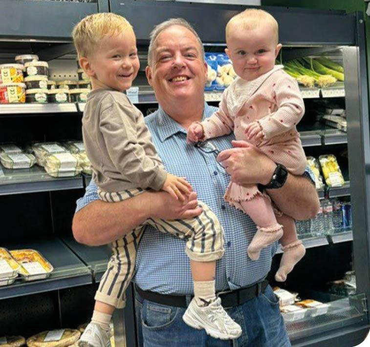 An adult wearing a blue checkered shirt and jeans is smiling while holding two children, one dressed in a beige outfit with striped pants and the other in a pink outfit, inside a store with refrigerated shelves. Description generated by AI