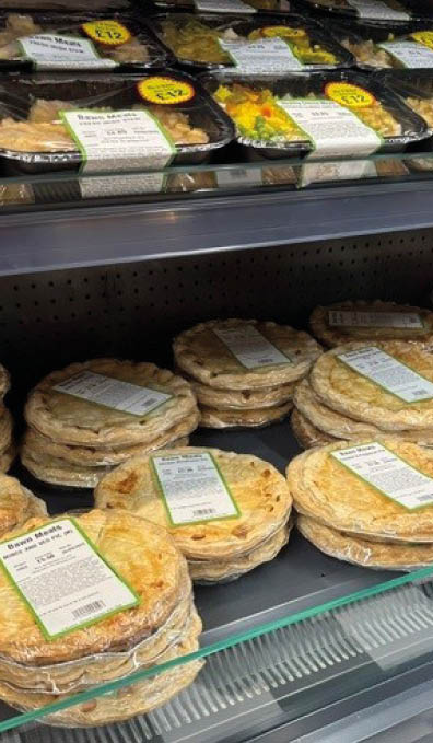 Stacks of packaged pies and ready-to-eat meals are displayed on glass shelves in a refrigerated store case. Description generated by AI