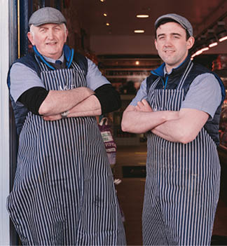 Two adults wearing striped aprons and flat caps stand with arms crossed at the entrance of a shop. Description generated by AI