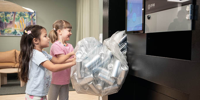 Two children with long hair and casual clothing are placing a large bag filled with aluminum cans into a recycling machine in a room with a couch and a painting on the wall. Description generated by AI