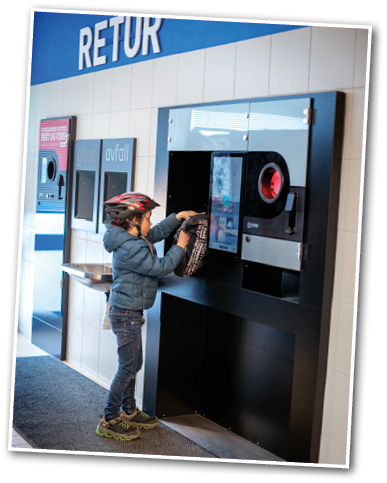 A child wearing a helmet and jacket is placing a bag into a reverse vending machine labeled “RETUR" in a clean indoor recycling area. Description generated by AI