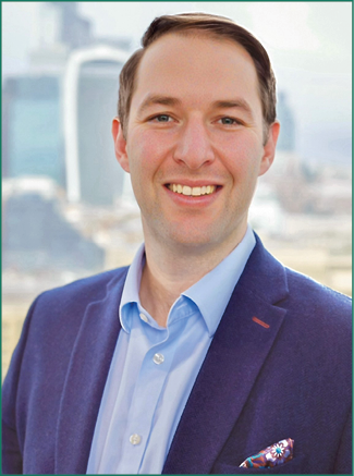 A person with short hair wearing a blue blazer and light blue shirt is smiling with a cityscape blurred in the background. Description generated by AI