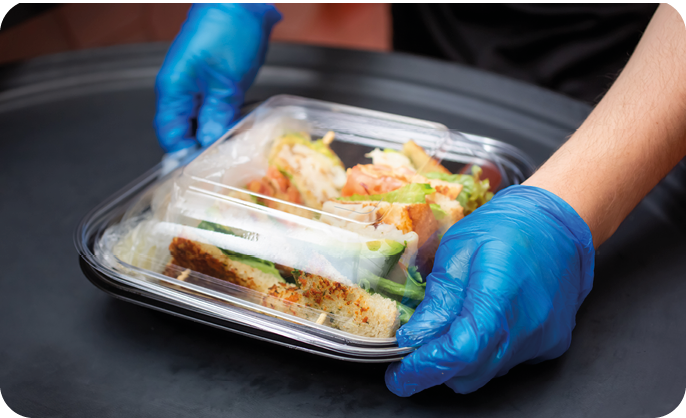 A person wearing blue gloves is holding a clear plastic container with a sandwich and other food items inside. Description generated by AI