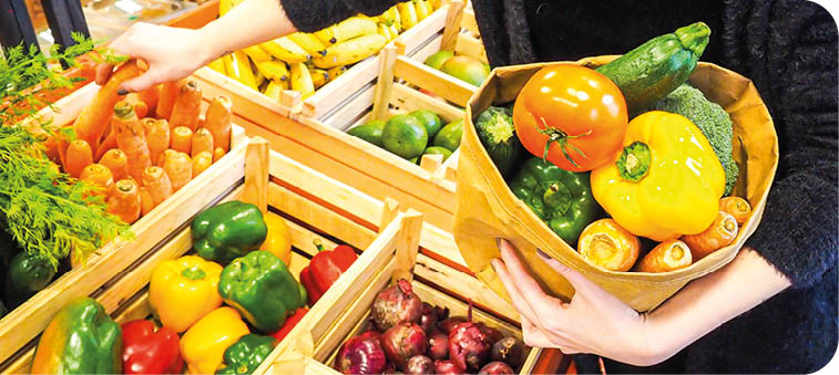 A person wearing a dark sweater is selecting fresh vegetables including carrots, bell peppers, onions, and zucchini from wooden crates at a market. Description generated by AI