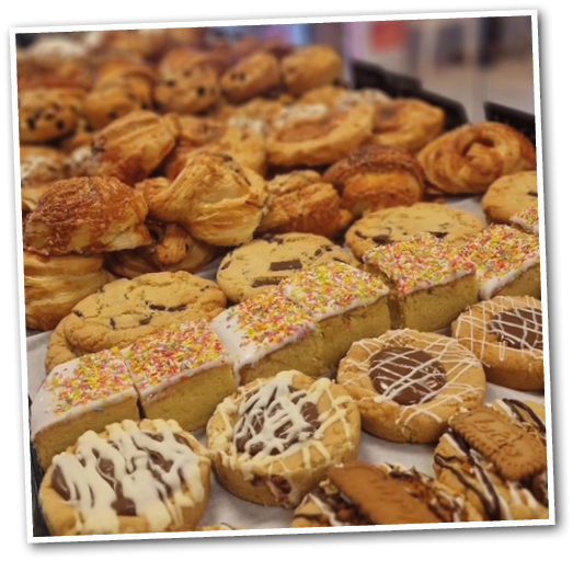 A variety of baked goods including cookies with chocolate chips, frosted cake squares with colorful sprinkles, and cookies decorated with white and chocolate drizzle are displayed on a tray in a bakery setting. Description generated by AI