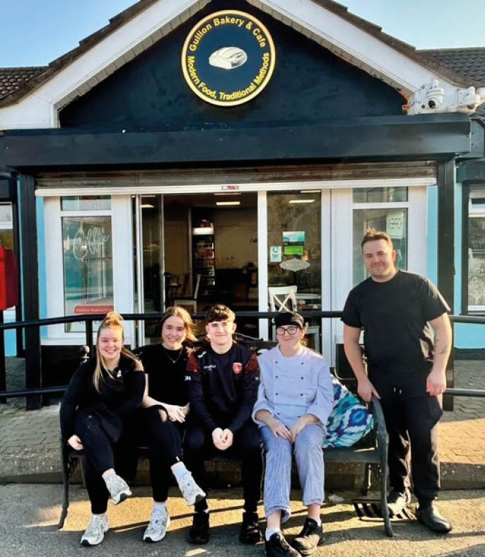 Five people dressed in casual and work attire are sitting and standing on a bench outside a bakery and caf on a sunny day.  Description generated by AI