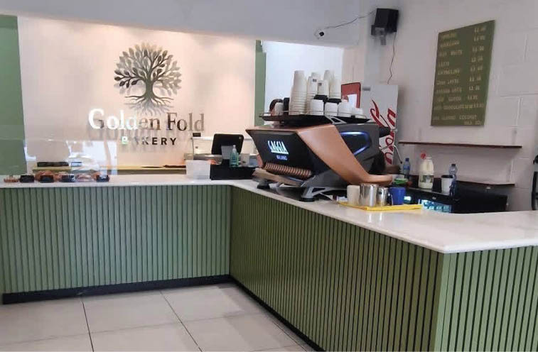 A modern bakery counter with green paneling and a person working behind it in a clean, well-lit interior with a tree logo and the words “Golden Fold Bakery" on the back wall. Description generated by AI