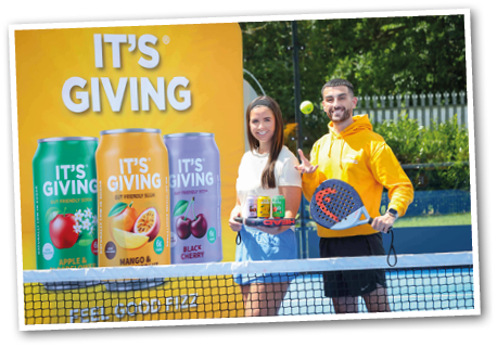 Two adults stand on a tennis court smiling with one holding a tray of canned drinks and the other holding a paddle and tossing a ball, in front of a large colorful advertisement for fruit-flavored sparkling water. Description generated by AI