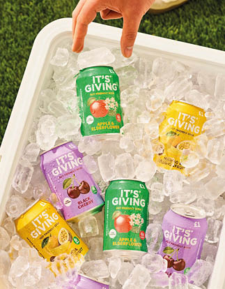 A hand is reaching into a cooler filled with ice and colorful cans labeled “It's Giving" with various fruit flavors on a grassy surface. Description generated by AI