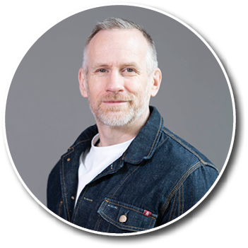 A person with short light hair and a beard is wearing a dark denim jacket over a white shirt and smiling slightly against a plain gray background. Description generated by AI